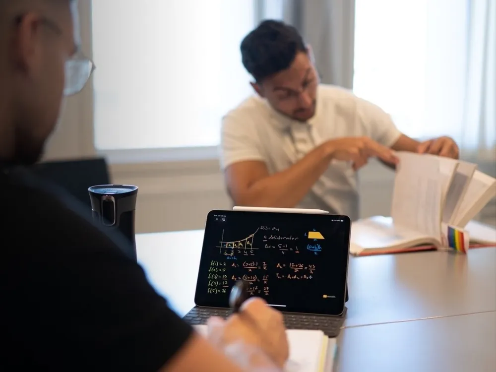 To personer jobber med matematikk ved et bord. Den ene noterer i kladdeboka ved siden av et nettbrett som viser en håndskrevet utregning av arealet under en graf, og den andre blar i et kompendium.